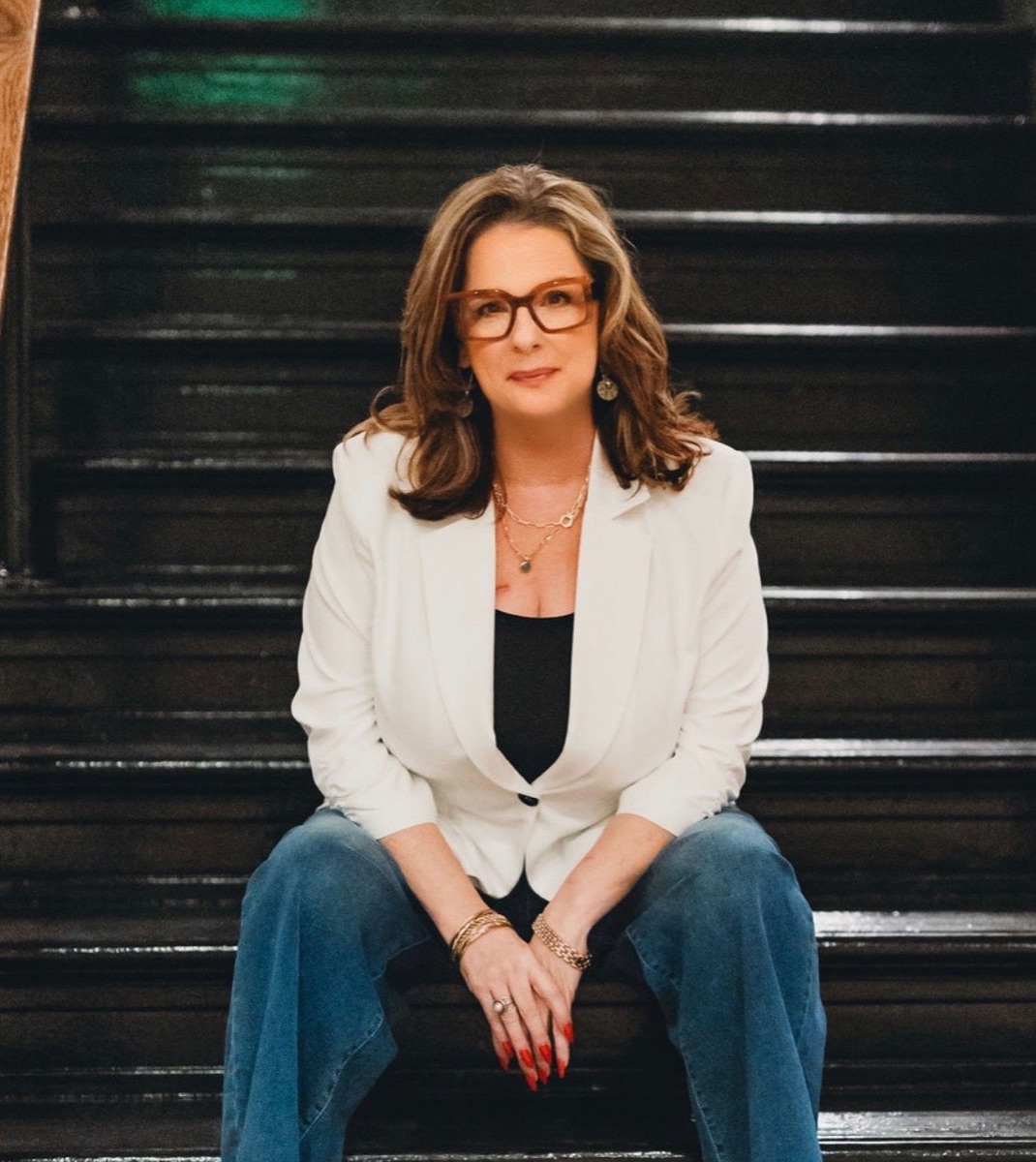 Allison Bush seated on steps in a white blazer and glasses