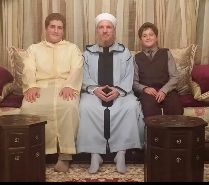 A white-bearded teacher in a white cap and a pale blue robe sits at the centre of a sofa, hands folded, between two young men — one in a pale yellow…